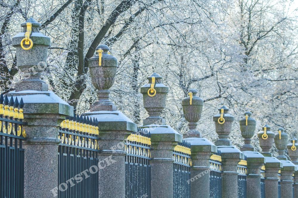 На снимке изображена гранитная ограда Летнего сада в Санкт-Петербурге. Золотые детали декора контрастируют с белоснежным налётом и искрящимися деревьями. Эта ограда — яркий пример элегантности и стиля петербургской архитектуры. Зима придаёт ансамблю сказочный вид, подчёркивая гармонию искусственного и природного.